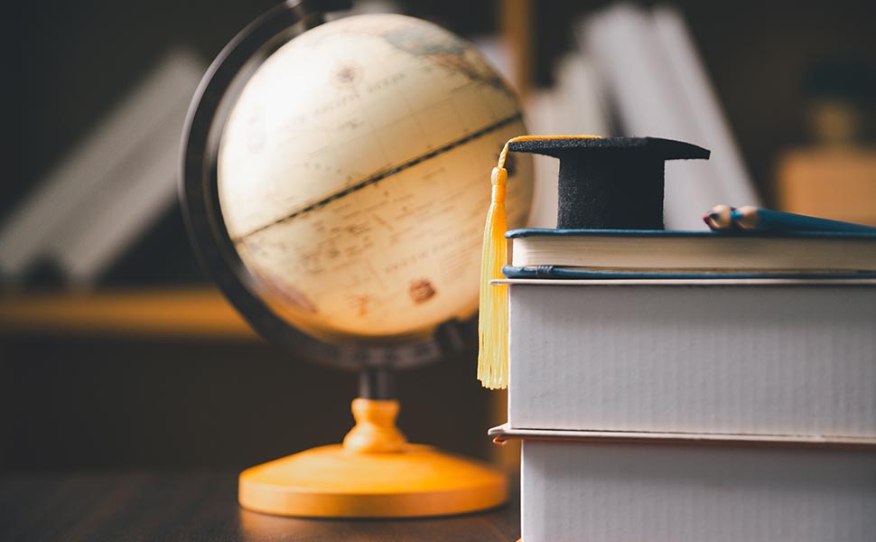 Graduation cap placed on stacked books beside a large classroom globe, representing education, geography, global learning, academic achievement, knowledge, study, scholarship, and school success.