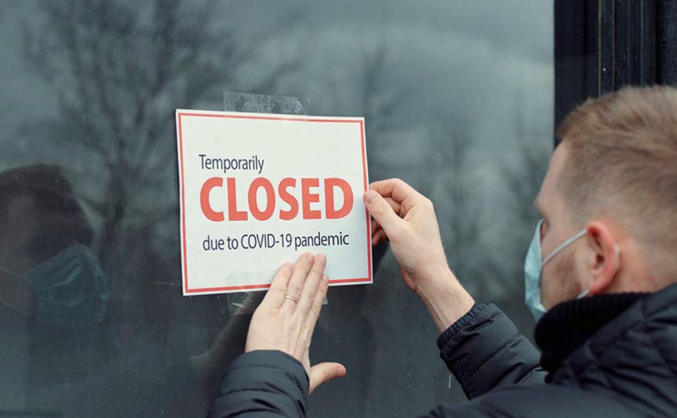 Caucasian male wearing medical mask puts a Temporary closed due COVID-19 pandemic sign on a window. Coronavirus pandemic, small business shutdown